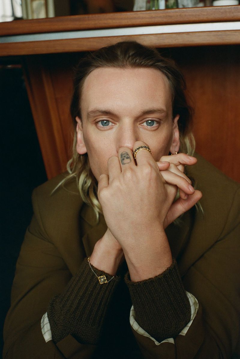 Close-up portrait of fair-skinned male with blonde hair, wearing green blazer and gold rings, his hands folded in front of his face.