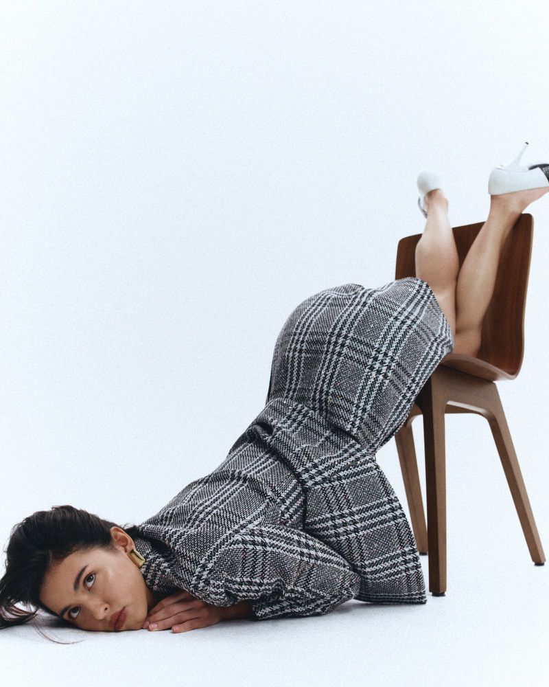 Woman in a grey plaid blazer and white heels poses on the floor with her legs resting on a brown chair in a bright photo studio.