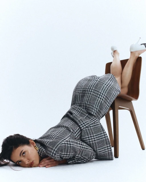 Woman in a grey plaid blazer and white heels poses on the floor with her legs resting on a brown chair in a bright photo studio.