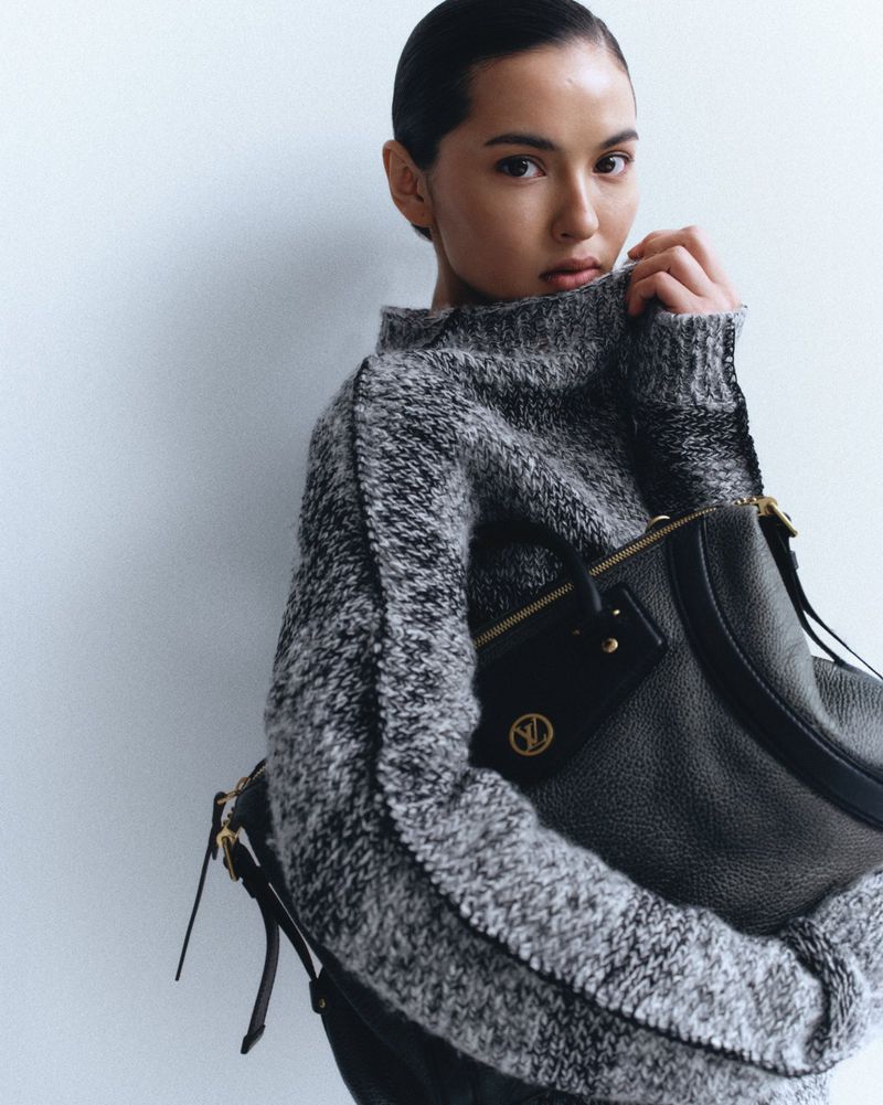 Woman in grey sweater holding a black leather purse with gold hardware against a white background in a studio-style shot.