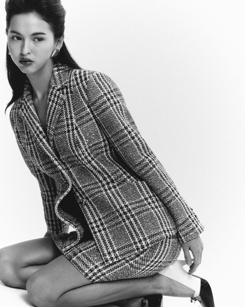 Black and white fashion portrait of a woman in a plaid blazer and skirt, kneeling in a photo studio with soft, diffused lighting.