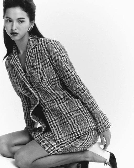 Black and white fashion portrait of a woman in a plaid blazer and skirt, kneeling in a photo studio with soft, diffused lighting.