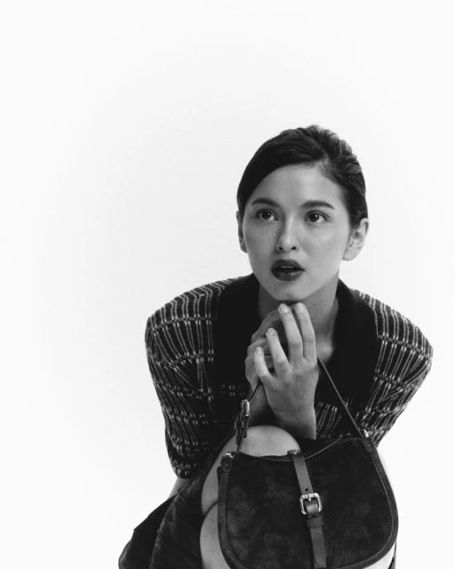 Monochrome portrait of a young woman in a patterned top and handbag, hands clasped beneath her chin, looking off-camera against a white backdrop.