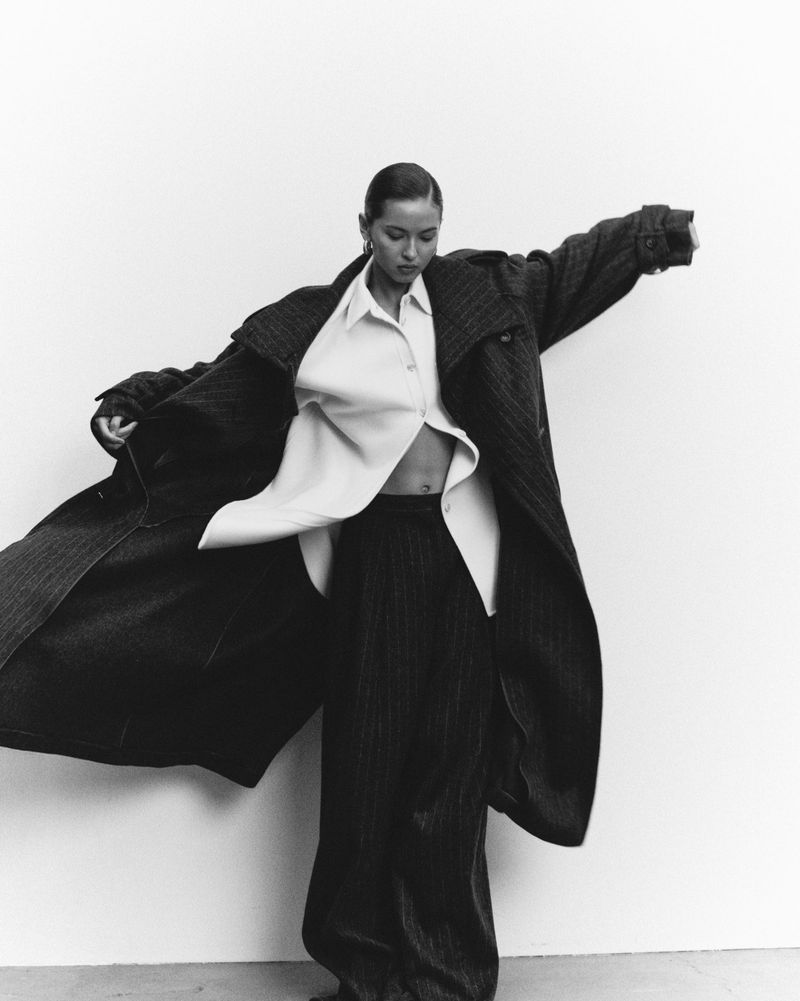 A woman wears a long pinstripe coat and matching wide-leg trousers over an unbuttoned white shirt, posing against a white studio wall.