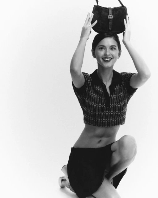 Woman kneeling in a photo studio holding a black bag over her head. The woman wears a polo shirt and a skirt, in black and white.