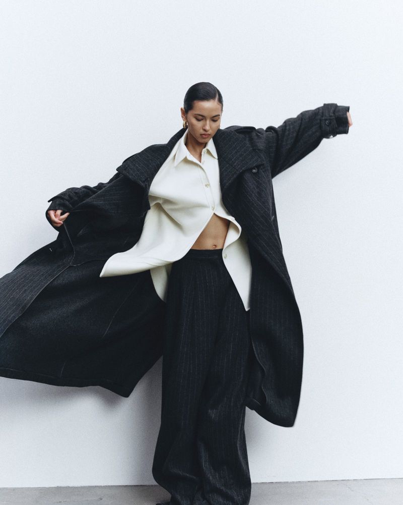 Editorial fashion shot of a woman in a beige shirt, pinstripe pants, and oversized coat against a neutral backdrop, arms extended.