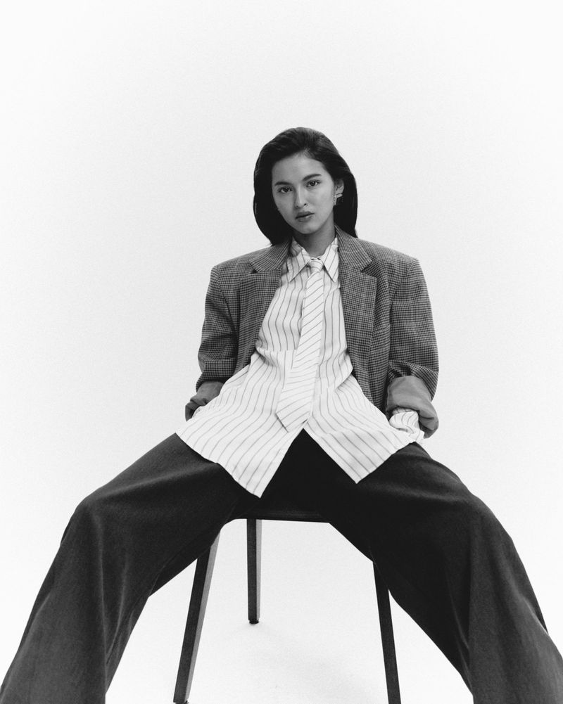 Woman in an oversized plaid blazer, striped shirt, and tie sitting on a chair against a white background