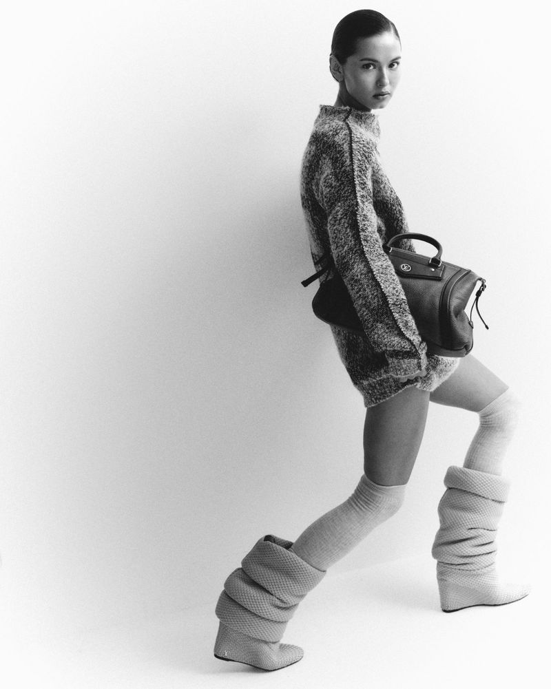 Fashion shot of a woman with slicked-back hair wearing a sweater dress, thigh-high socks, and chunky heeled boots, carrying a handbag.
