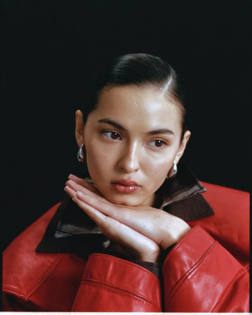 Portrait of a woman with dark hair wearing a red leather jacket, resting her chin on her hands against a dark background.