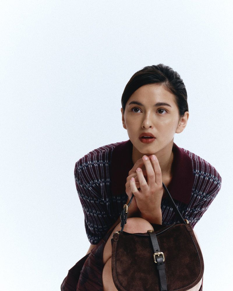 Woman in a plaid dress and brown bag, with hands near her face, gazes upwards against a white background, creating a studio portrait.