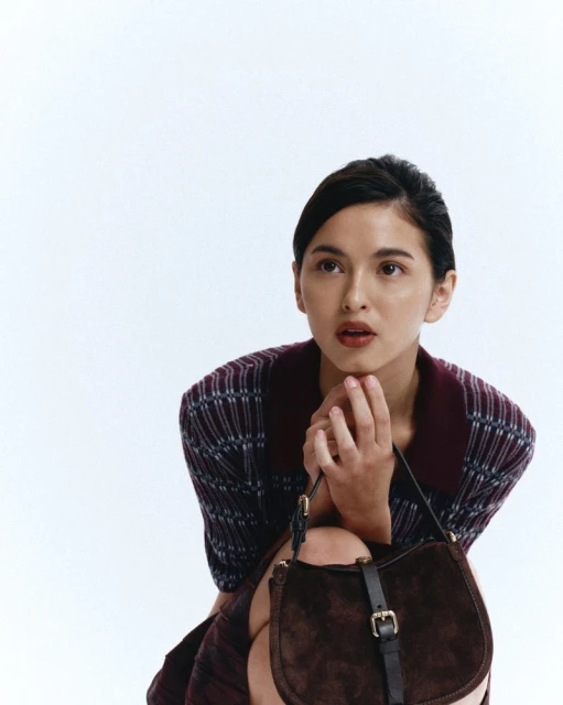 Woman in a plaid dress and brown bag, with hands near her face, gazes upwards against a white background, creating a studio portrait.
