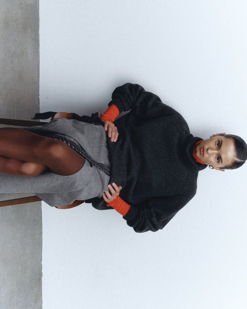 A woman is sitting on a chair, wearing a gray sweater and skirt. The background is a plain white wall with soft, diffused lighting.