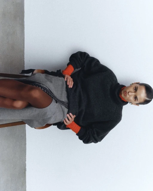 A woman is sitting on a chair, wearing a gray sweater and skirt. The background is a plain white wall with soft, diffused lighting.