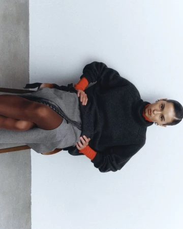 A woman is sitting on a chair, wearing a gray sweater and skirt. The background is a plain white wall with soft, diffused lighting.