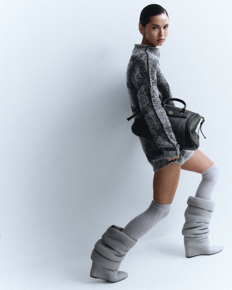 A stylish woman poses in a grey knit sweater dress and thigh-high knit boots, accessorized with a black leather satchel against a white backdrop.
