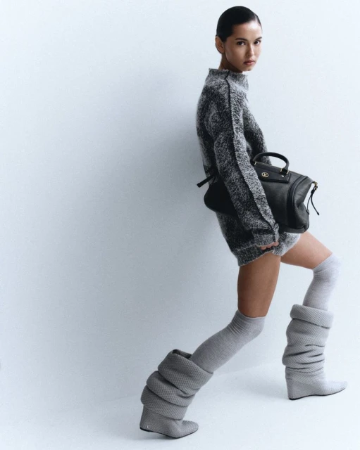 A stylish woman poses in a grey knit sweater dress and thigh-high knit boots, accessorized with a black leather satchel against a white backdrop.