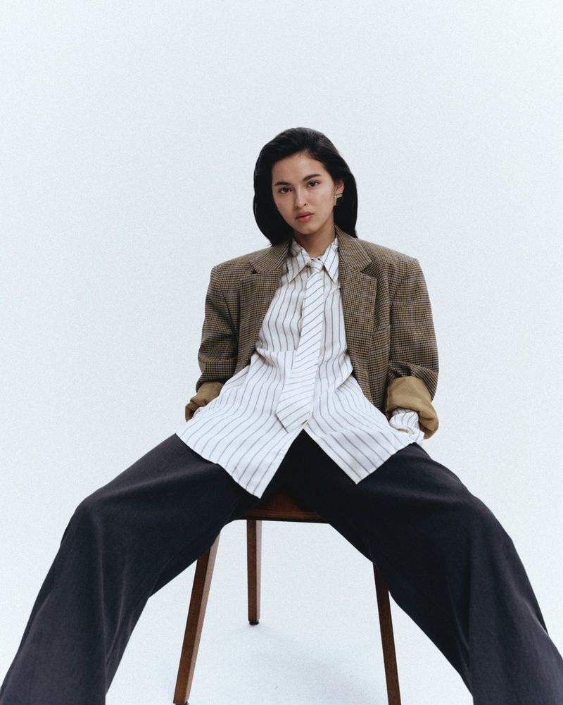 Woman seated on a chair wearing a brown plaid blazer, striped shirt, tie, and dark trousers against a white seamless background