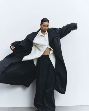 Model in a long dark coat and white shirt posing against a plain white background for an editorial spread
