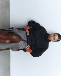 Model seated on a chair wearing a dark grey turtleneck sweater and grey skirt against a white studio background.