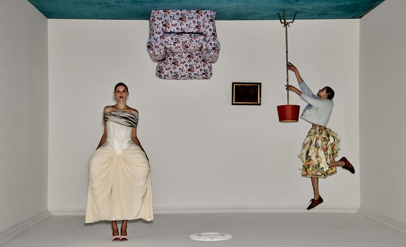 Fashion photo in a surreal, inverted room with two women, an armchair on the ceiling, and a bucket hanging from the ceiling.