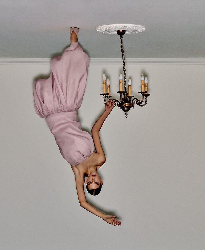 Upside-down view of a woman in a pink dress, her feet on the ceiling, hanging from an ornate chandelier in a white room.
