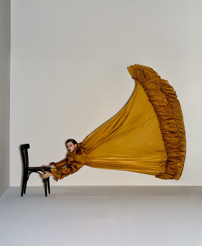 A woman in a gold gown stretches horizontally between a black chair and a neutral background in a studio, showcasing a unique fashion pose.
