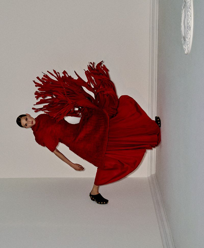 Fashion shot of woman in red dress and black clogs striking a pose in a corner against a white wall and floor in a brightly lit room.