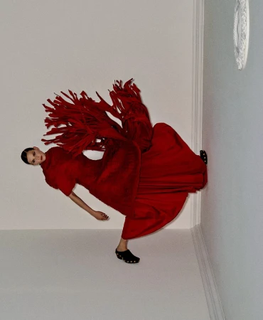 Fashion shot of woman in red dress and black clogs striking a pose in a corner against a white wall and floor in a brightly lit room.
