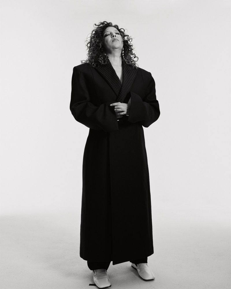 Adult woman with curly hair wearing a black overcoat and cream colored flats stands against a white background in a studio shot.
