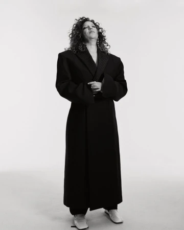 Adult woman with curly hair wearing a black overcoat and cream colored flats stands against a white background in a studio shot.
