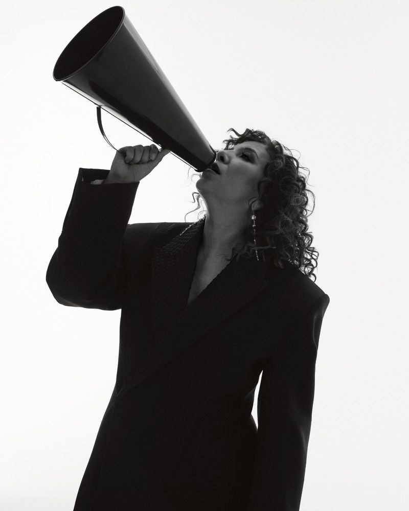 Monochromatic portrait of a curly-haired woman in a blazer speaking into a large, black megaphone against a white backdrop.