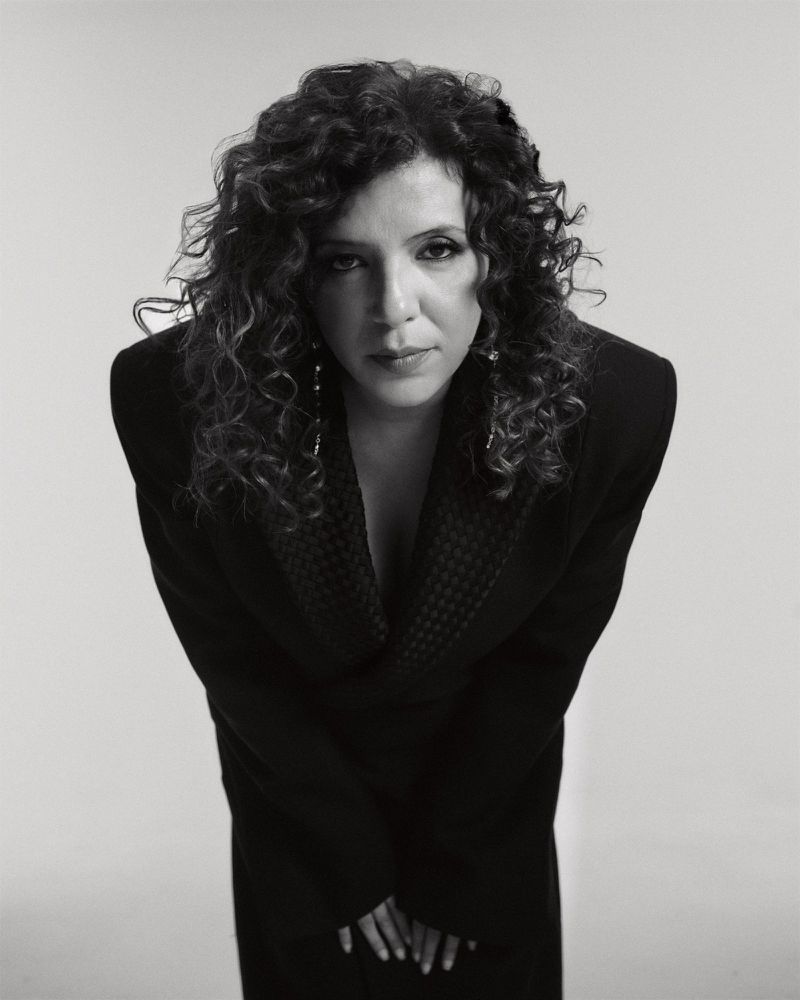 Black-and-white portrait of an adult woman with curly hair, wearing a black jacket in a studio setting, captured at an eye-level angle.