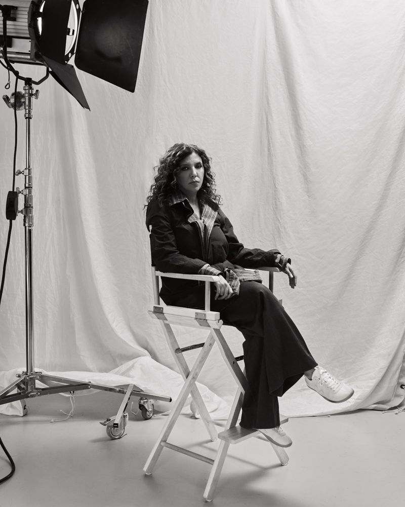 Black and white portrait of a woman seated on a director's chair in a photo studio with a light stand and backdrop.