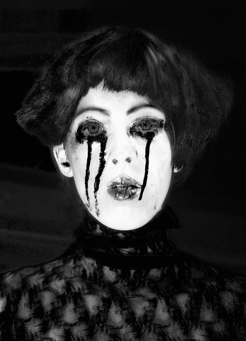 Striking black and white portrait of a woman with dark eye makeup, a lace top, and a retro hairstyle against a dark background.