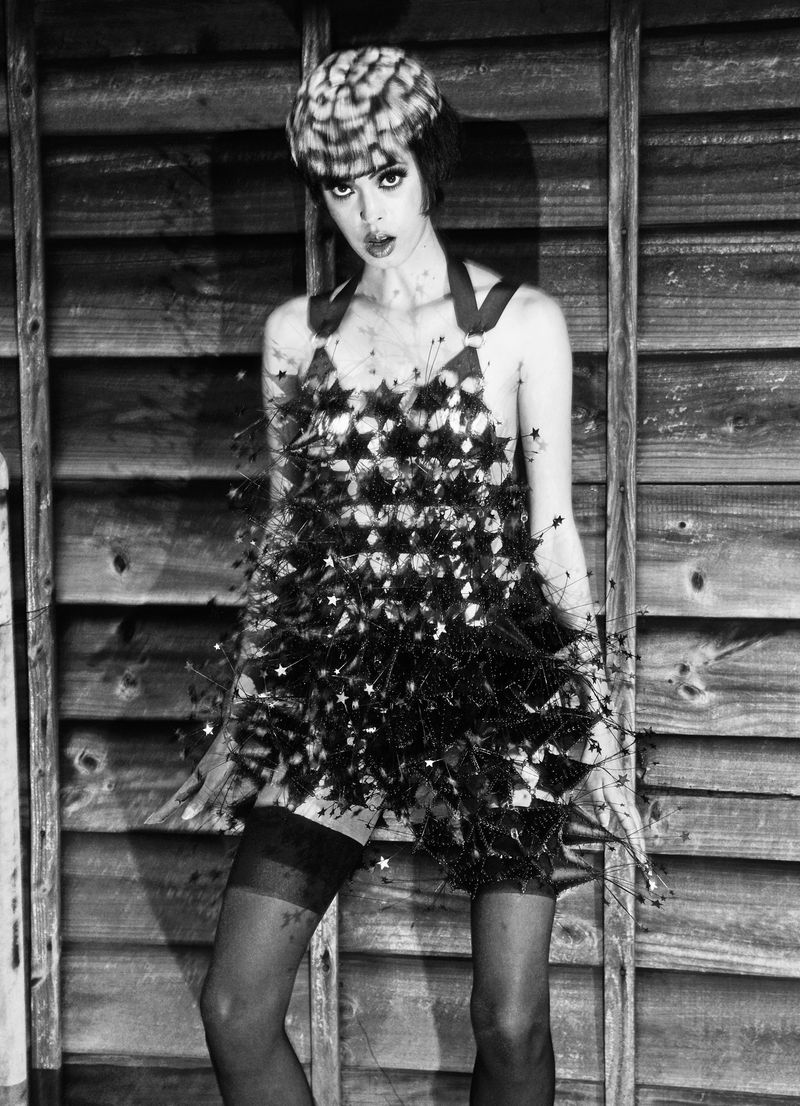 Monochrome portrait of a woman wearing a textured hat and star-adorned dress, posing against a weathered wooden wall in a fashion setting.