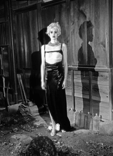 Monochrome portrait of a woman with platinum blonde hair wearing a black dress and jewelry, standing in front of a wooden wall and bottles.