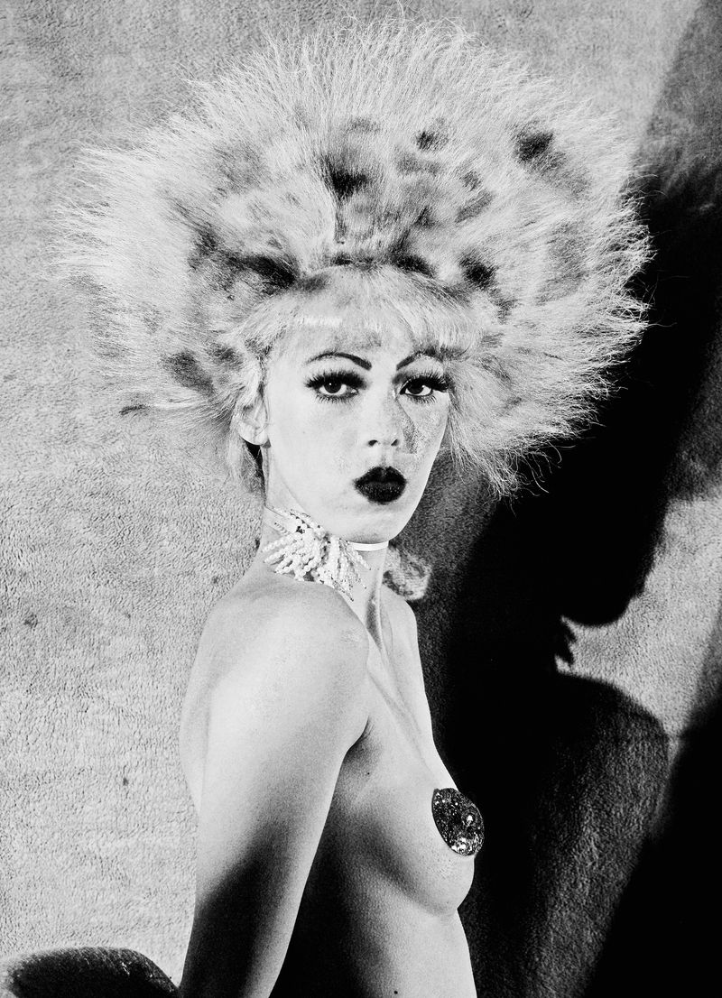 Striking portrait of a fair-skinned person with elaborate feathered headwear, dark lipstick, and decorative body jewelry against a textured backdrop.