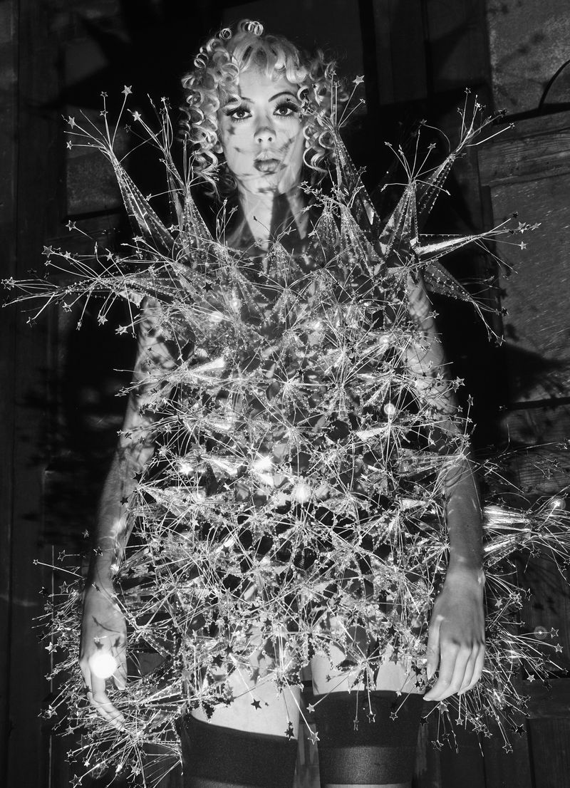 Black and white shot of a person wearing an elaborate, reflective costume with spiky, starburst-like elements, posing indoors.