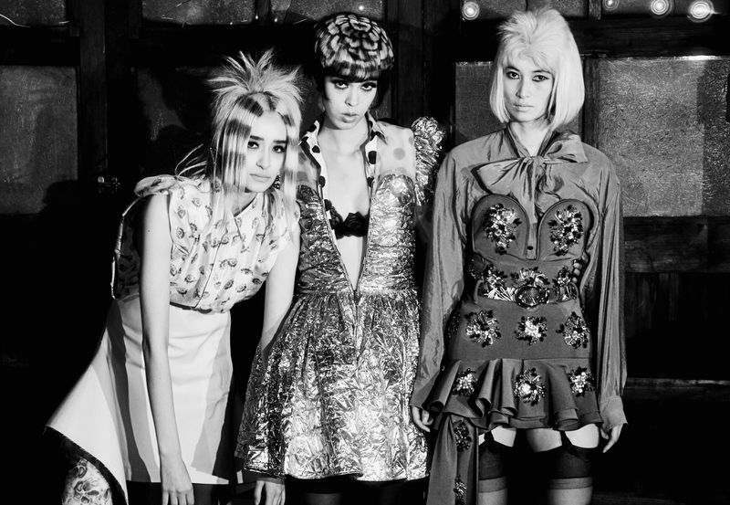 Three women in avant-garde fashion stand in a monochromatic indoor setting, showcasing unique hairstyles and elaborate dresses with detailed textures.