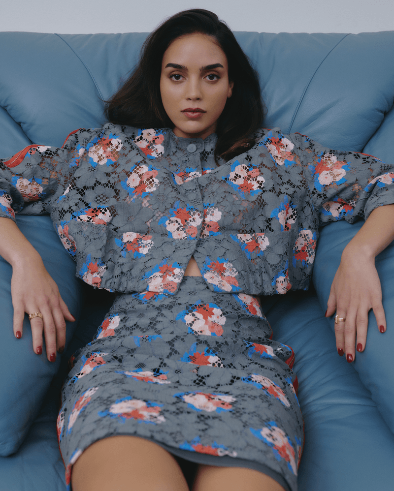 A woman poses on a blue leather couch wearing a floral-patterned matching skirt and jacket. Her dark hair frames her face and she has a neutral expression.