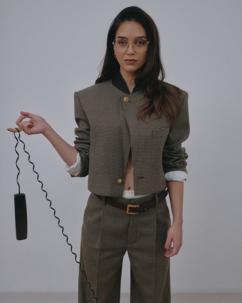 A woman in a houndstooth blazer and pants holds a black phone receiver with cord against a neutral backdrop, gazing at the camera.