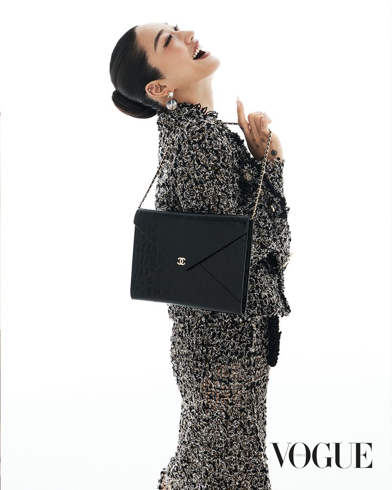 An Asian female fashion model in a tweed skirt suit poses in a photo studio holding a black Chanel clutch purse with gold logo hardware.