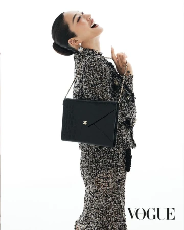 An Asian female fashion model in a tweed skirt suit poses in a photo studio holding a black Chanel clutch purse with gold logo hardware.