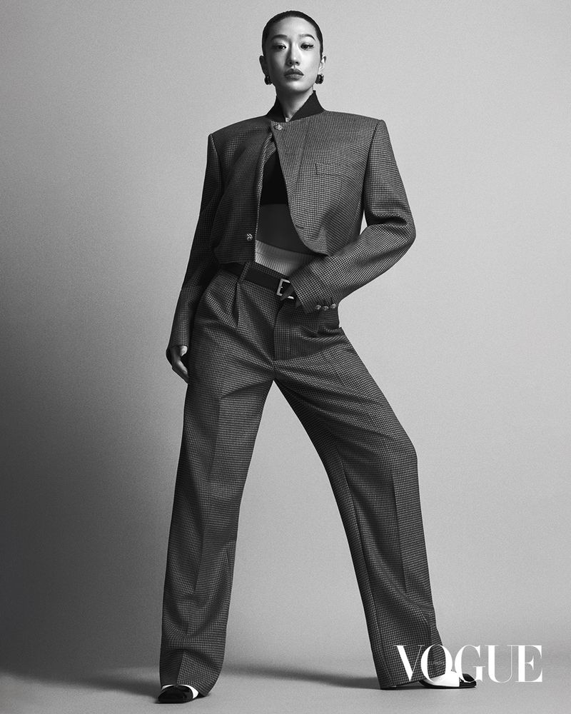 An adult woman with short dark hair poses in a grey patterned cropped jacket and matching pants, highlighted by studio lighting on a plain backdrop.