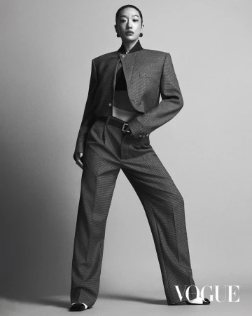 An adult woman with short dark hair poses in a grey patterned cropped jacket and matching pants, highlighted by studio lighting on a plain backdrop.