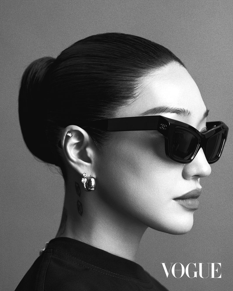 Black and white close-up of a woman with dark hair in a bun wearing oversized sunglasses, hoop earrings, and a black top.