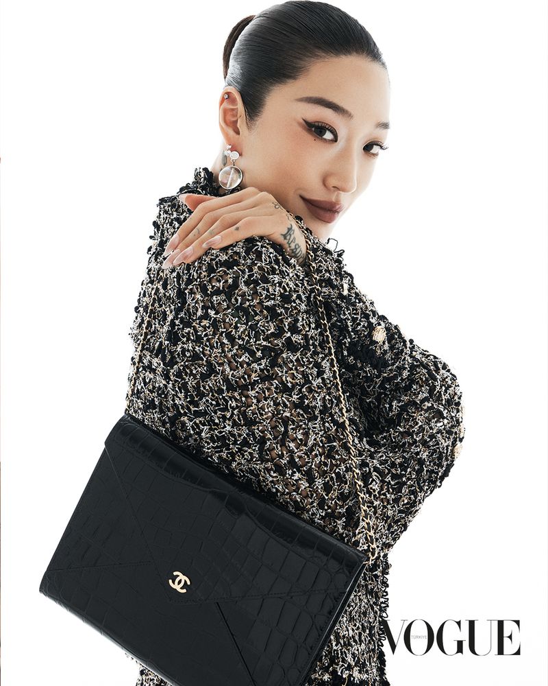 Model wearing a black tweed jacket and holding a black leather handbag against a bright white background