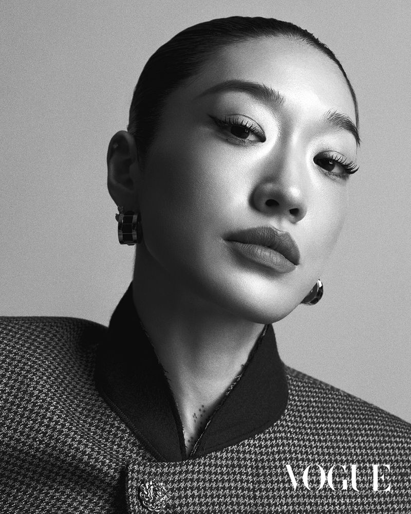 Headshot of a woman with slicked-back hair, featuring a houndstooth blazer, statement earrings, and bold winged eyeliner, in black and white.