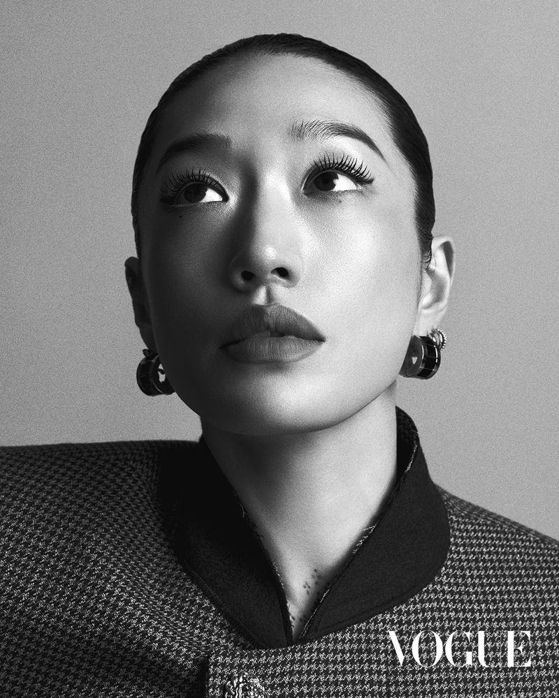 Headshot of a stylish Asian woman with makeup and jewelry, looking up. She is wearing a patterned suit jacket in a studio setting.
