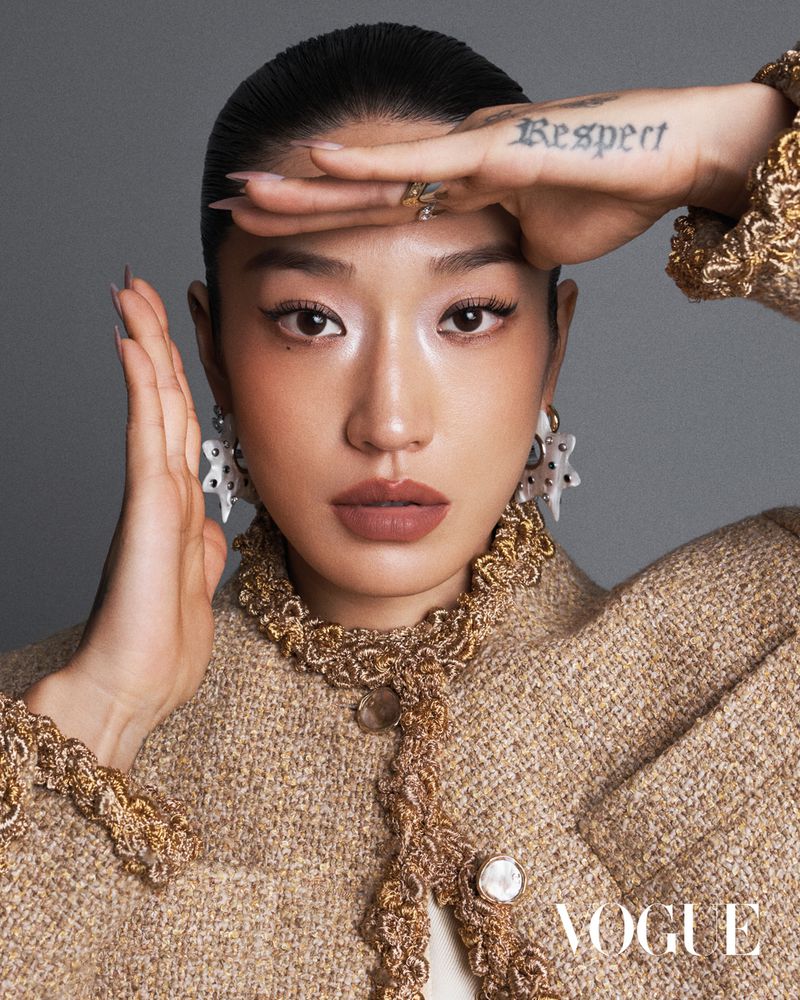 Portrait of Asian woman wearing tweed jacket and starburst earrings, shielding eyes with tattooed hand, with Vogue logo at the bottom.